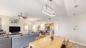 A modern living room with a sofa, TV, and dining area with a wooden table and chairs. - Spacious condo with bay views, renovated pool, hot tub, & beach access! (Miramar Beach)