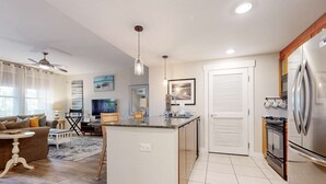 A modern kitchen with a central island, a dining area with a sofa and chairs, and a living room with a TV and a coffee table. - Spacious condo with bay views, renovated pool, hot tub, & beach access! (Miramar Beach)
