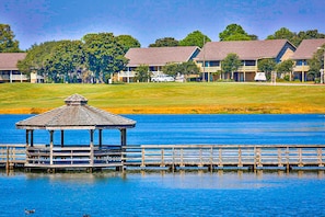 Ein Pavillon auf einem Steg über dem Wasser, mit einem Golfplatz und Gebäuden im Hintergrund.