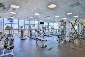 Ein Fitnessstudio mit unterschiedlichen Trainingsgeräten, großen Fenstern und einem Deckenventilator.