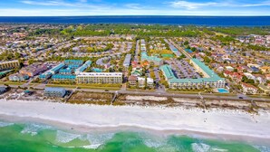 Vue aérienne d’une ville côtière avec un hôtel en bord de mer, comprenant un grand espace piscine, plusieurs bâtiments et une vue dégagée sur l’océan.