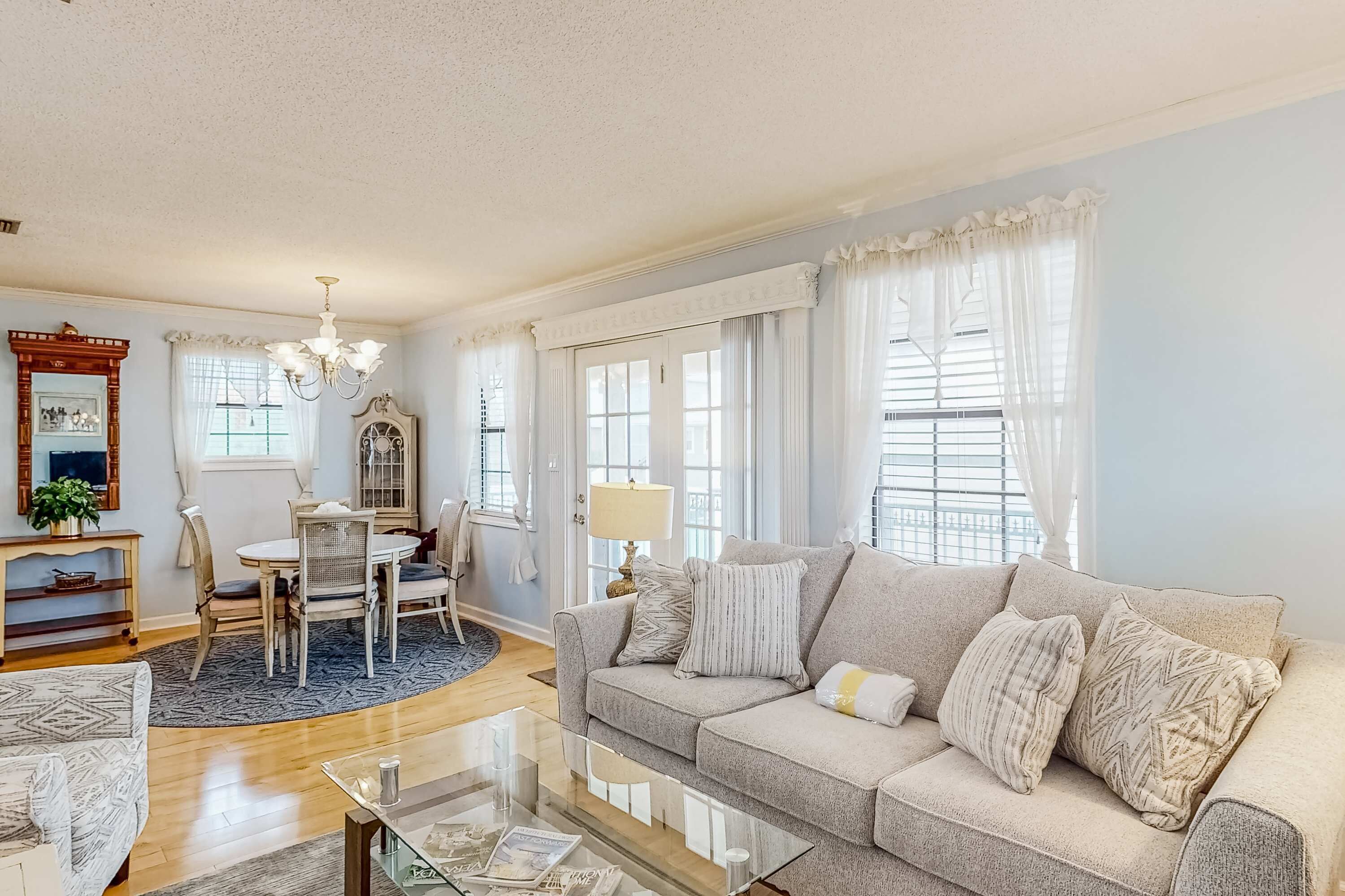 A living room with a beige sofa, a glass coffee table, a dining table with chairs, and a chandelier.
