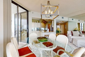 A modern living room with a glass dining table, white chairs, a sofa, and a chandelier. - Beautiful beachfront condo w/ shared pool, tennis, & beach access (Miramar Beach)