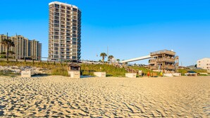 Ein sandiger Strand mit einem hohen Gebäude und einer Holzkonstruktion im Hintergrund.