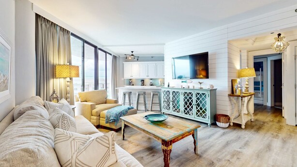 A modern living room with a sofa, a coffee table, and a kitchen area with white cabinets and a TV. - Cheerful 10th Floor Condo with Shared Pool & Hot Tub, WiFi, and Central AC (Miramar Beach)