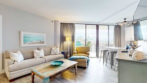 A modern living room with a sofa, armchair, and coffee table. There is a large window with curtains, a painting on the wall, and a view of buildings outside. - Cheerful 10th Floor Condo with Shared Pool & Hot Tub, WiFi, and Central AC (Miramar Beach)