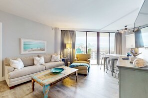 A modern living room with a sofa, armchair, and coffee table. There is a large window with curtains, a painting on the wall, and a view of buildings outside. - Cheerful 10th Floor Condo with Shared Pool & Hot Tub, WiFi, and Central AC (Miramar Beach)
