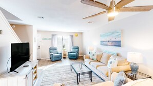 A modern living room with a flat-screen TV, two blue armchairs, a beige sofa, a glass coffee table, and a ceiling fan. - Beautifully updated 2nd-floor condo near the beach with pool, balcony, W/D, & AC (Miramar Beach)