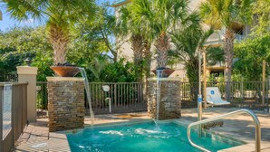 A swimming pool with a stone wall and a waterfall feature, surrounded by palm trees and a fence. - Beach service included at gated coastal condo with pool, hot tub, W&D, & balcony (Miramar Beach)