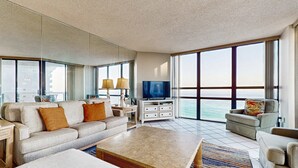 A spacious living room with a large flat-screen TV, a beige sofa, a coffee table, and a view of the ocean through large windows. - Spacious waterfront condo w/ shared pool/hot tub/gym & beach access (Miramar Beach)