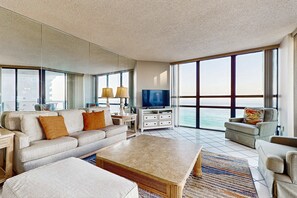A spacious living room with a large flat-screen TV, a beige sofa, a coffee table, and a view of the ocean through large windows. - Spacious waterfront condo w/ shared pool/hot tub/gym & beach access (Miramar Beach)