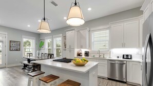 Cocina moderna con armarios blancos, una isla central con taburetes, electrodomésticos de acero inoxidable e iluminación colgante.
