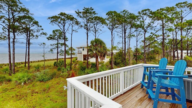 A wooden deck with blue Adirondack chairs overlooking a beach with tall pine trees and a white building in the background. - Gorgeous coastal home with plenty of space and easy beach access (St. George Island)