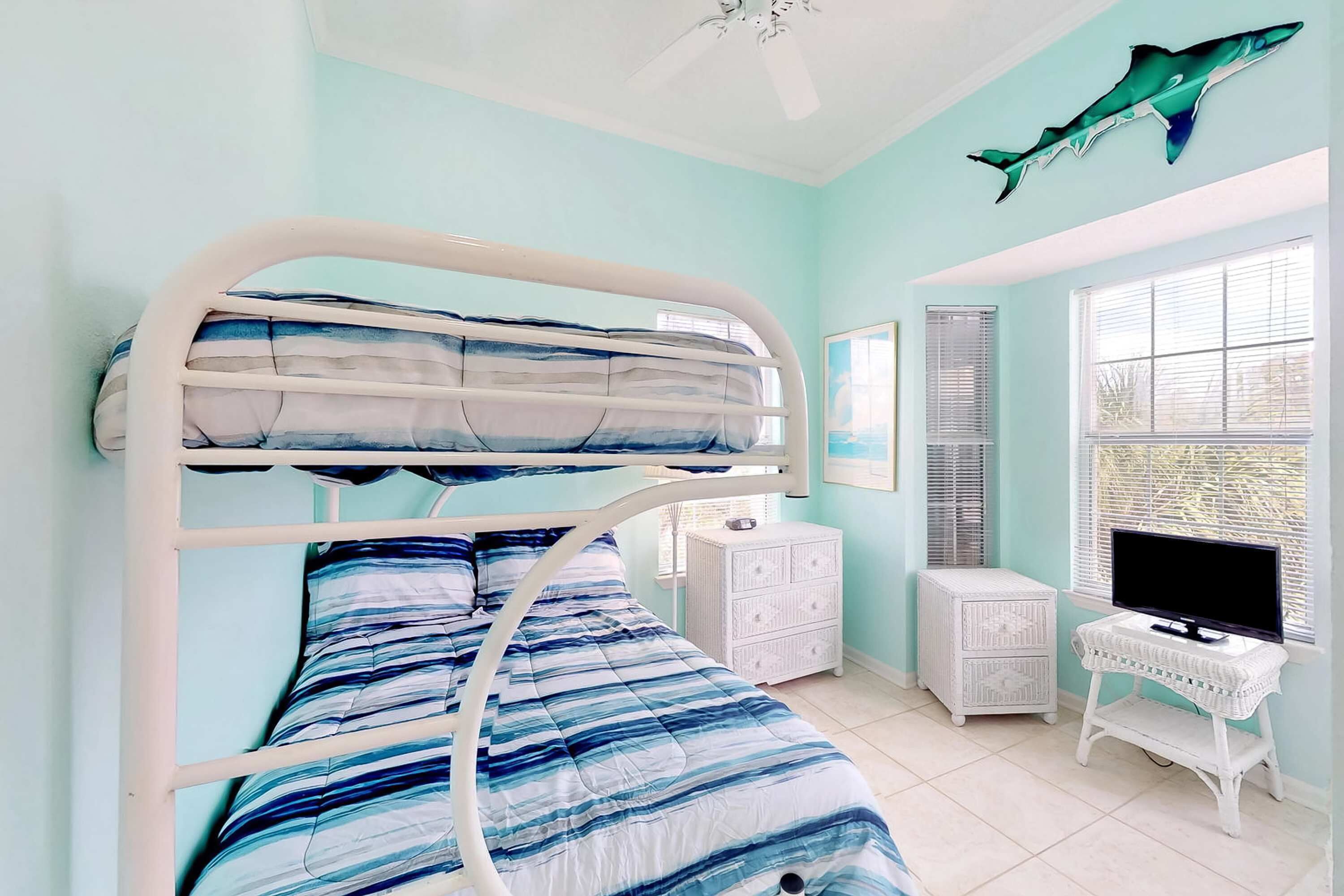 Un lit superposé avec une literie rayée bleue et blanche, une commode blanche, un téléviseur posé sur une petite table et une décoration représentant un requin sur le mur.