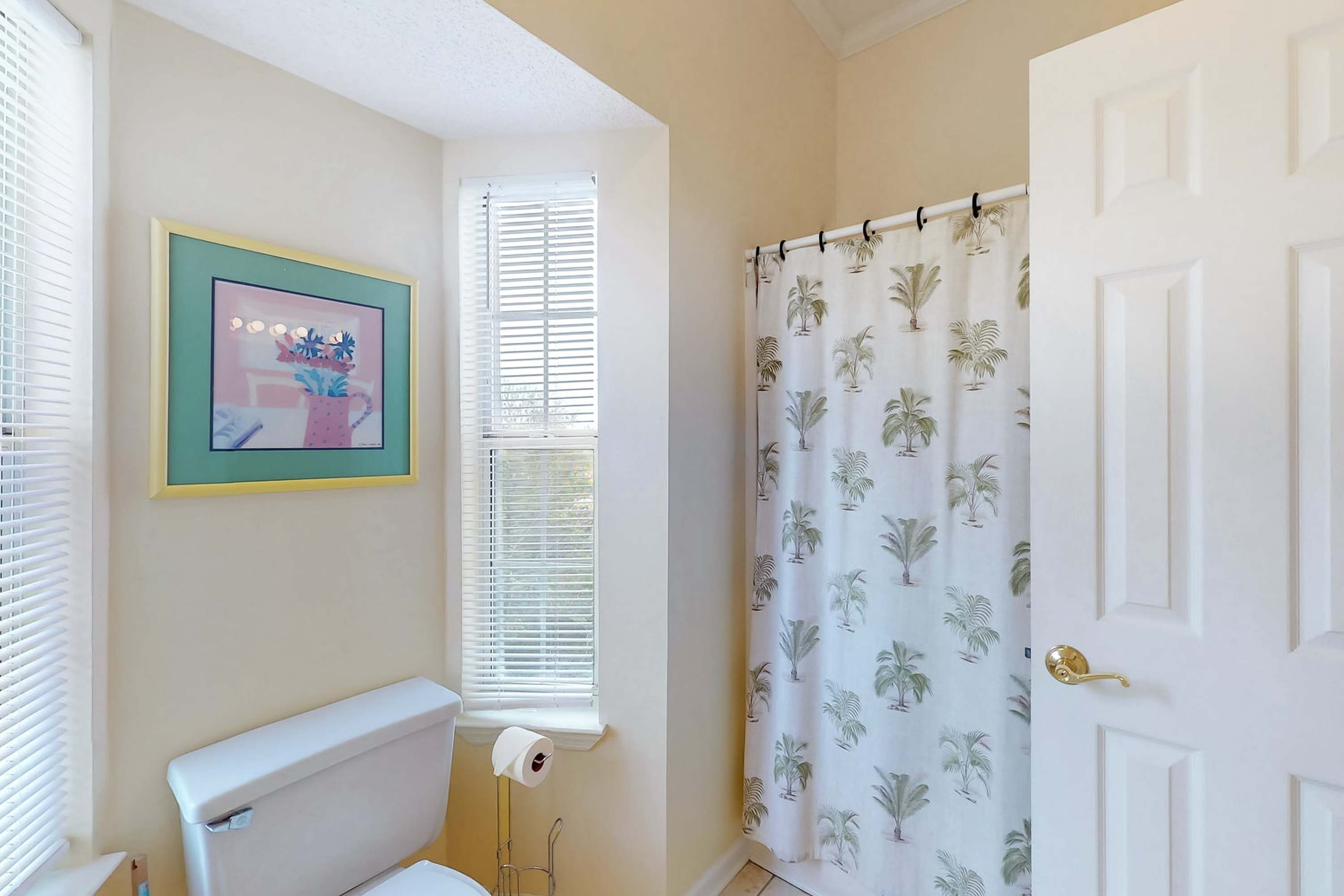 A bathroom with a toilet, a framed picture on the wall, a window with blinds, and a shower curtain with a palm leaf pattern.