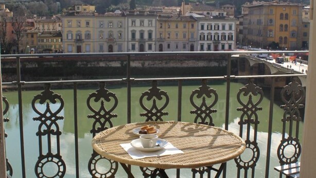 Uma varanda com mesa de vime, guarda-corpo e vista para um rio com edifícios ao fundo.