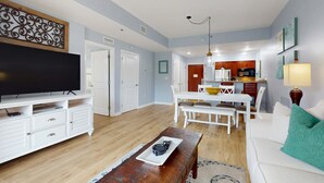 Een woonkamer met een televisie, witte meubels, een houten salontafel en een keuken op de achtergrond.