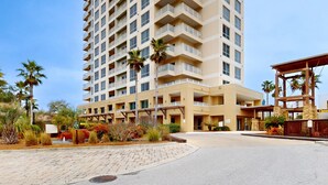 Een meerlaags hotel met balkons, palmbomen en een verhard entreegebied.