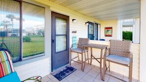 A covered porch with a sliding glass door, a small table with chairs, and a colorful striped chair. - Updated ground-floor condo with patio, W/D, pool, BBQ area, & AC - near beach (Miramar Beach)