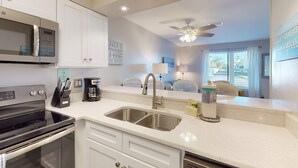 A modern kitchen with a microwave, stove, and sink. There is a ceiling fan and a view of a bedroom with a bed and window. - Updated ground-floor condo with patio, W/D, pool, BBQ area, & AC - near beach (Miramar Beach)