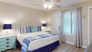 A bedroom with a bed, nightstands, a ceiling fan, a window with curtains, and a door with a cross on the wall. - Updated ground-floor condo with patio, W/D, pool, BBQ area, & AC - near beach (Miramar Beach)