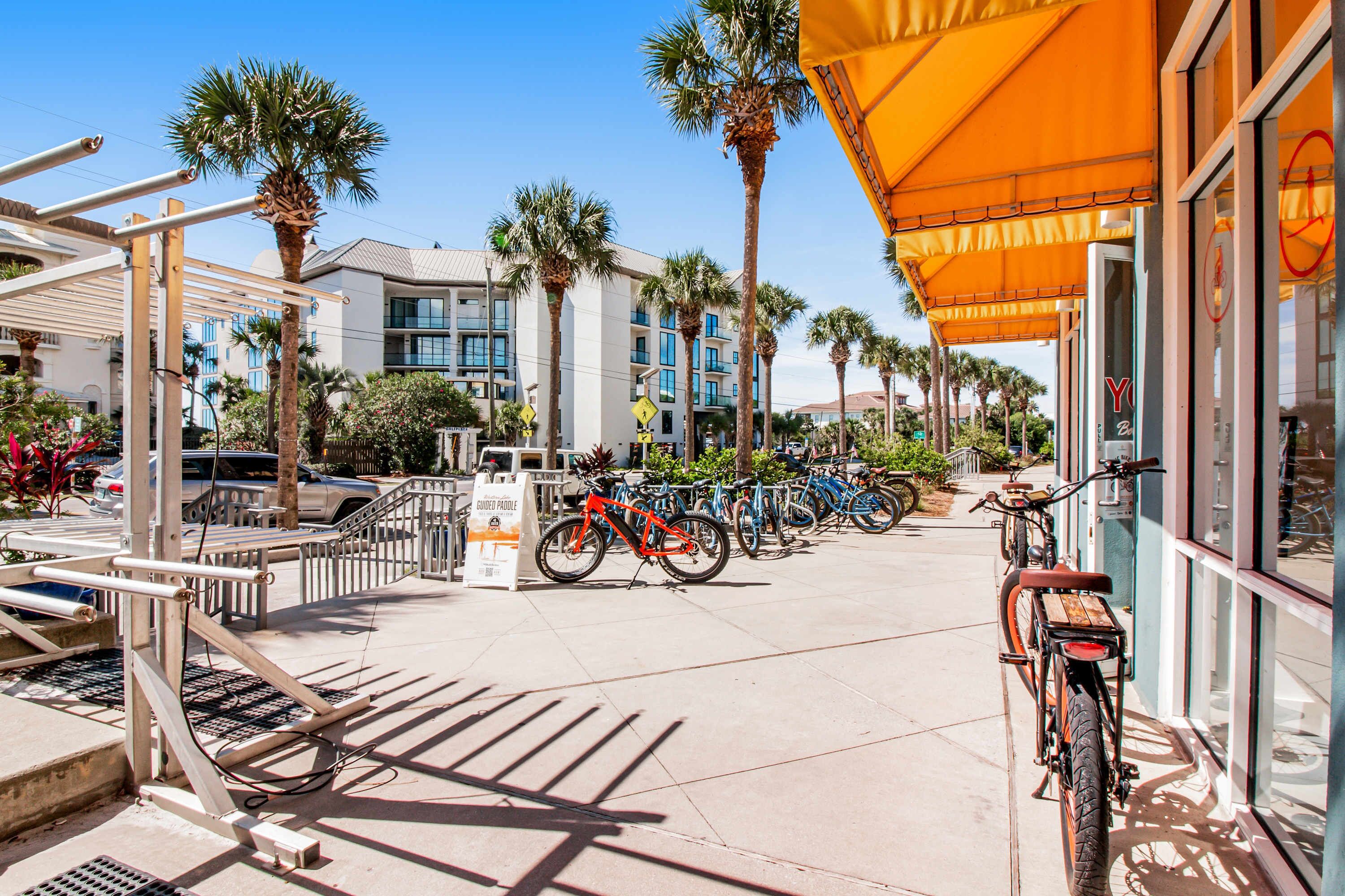A sunny street with bicycles parked along the sidewalk, palm trees, and a building with a yellow awning.