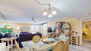 A spacious living room with a glass-top dining table, wicker chairs, a ceiling fan, and a television. - 1st floor comfortable Gulf view condo, beach setup included, near dining (Miramar Beach)