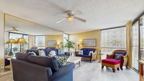 A spacious living room with a large sofa, a wicker chair, and a red ottoman. There is a ceiling fan, a large window with curtains, and a plant in the corner. - 1st floor comfortable Gulf view condo, beach setup included, near dining (Miramar Beach)