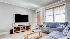 Una sala de estar con una televisión de pantalla plana, una mesa de centro de madera, un sofá gris y una ventana con persianas.