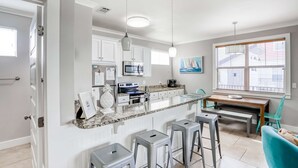 Una cocina moderna con armarios blancos, encimera de granito, horno empotrado y zona de comedor con mesa de madera y taburetes de bar.