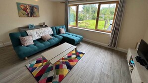 Ein Wohnzimmer mit einem salbeigreenen Sofa, einem Couchtisch, einem bunten Teppich, einem großen Fenster mit Vorhängen und einem Elefantenbild an der Wand.