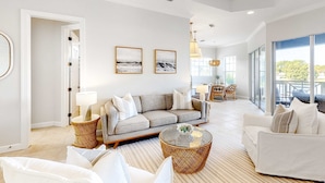 A spacious living room with a sofa, coffee table, and chairs, a dining area with a table and chairs, and large windows offering a view of the outdoors. - Lakefront condo with water-view balcony, private golf cart, resort pool & amenities (Miramar Beach)