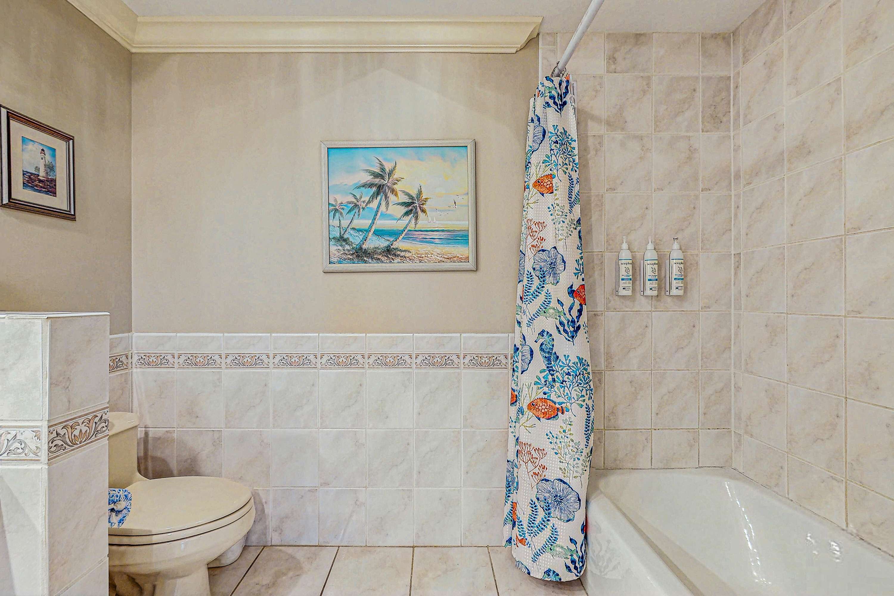 A bathroom with a toilet, a bathtub, and a shower curtain with a floral pattern. There is a framed picture on the wall above the toilet.