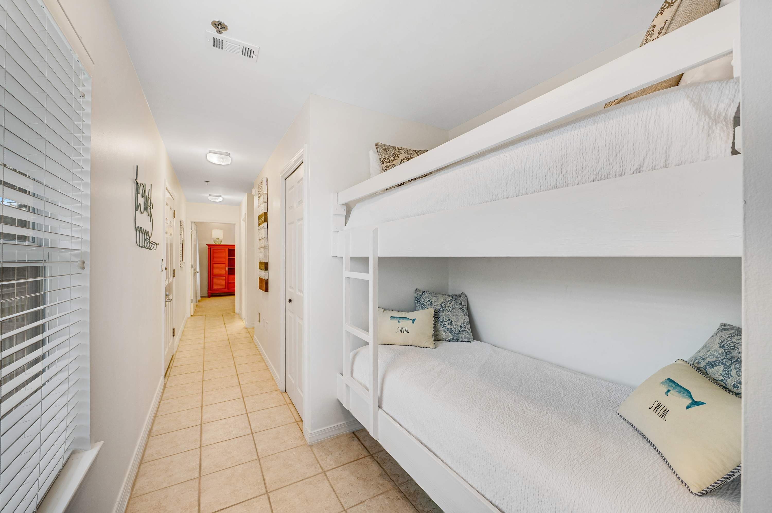 A narrow hallway with a bunk bed on the right, a red door at the end, and a window with blinds on the left.