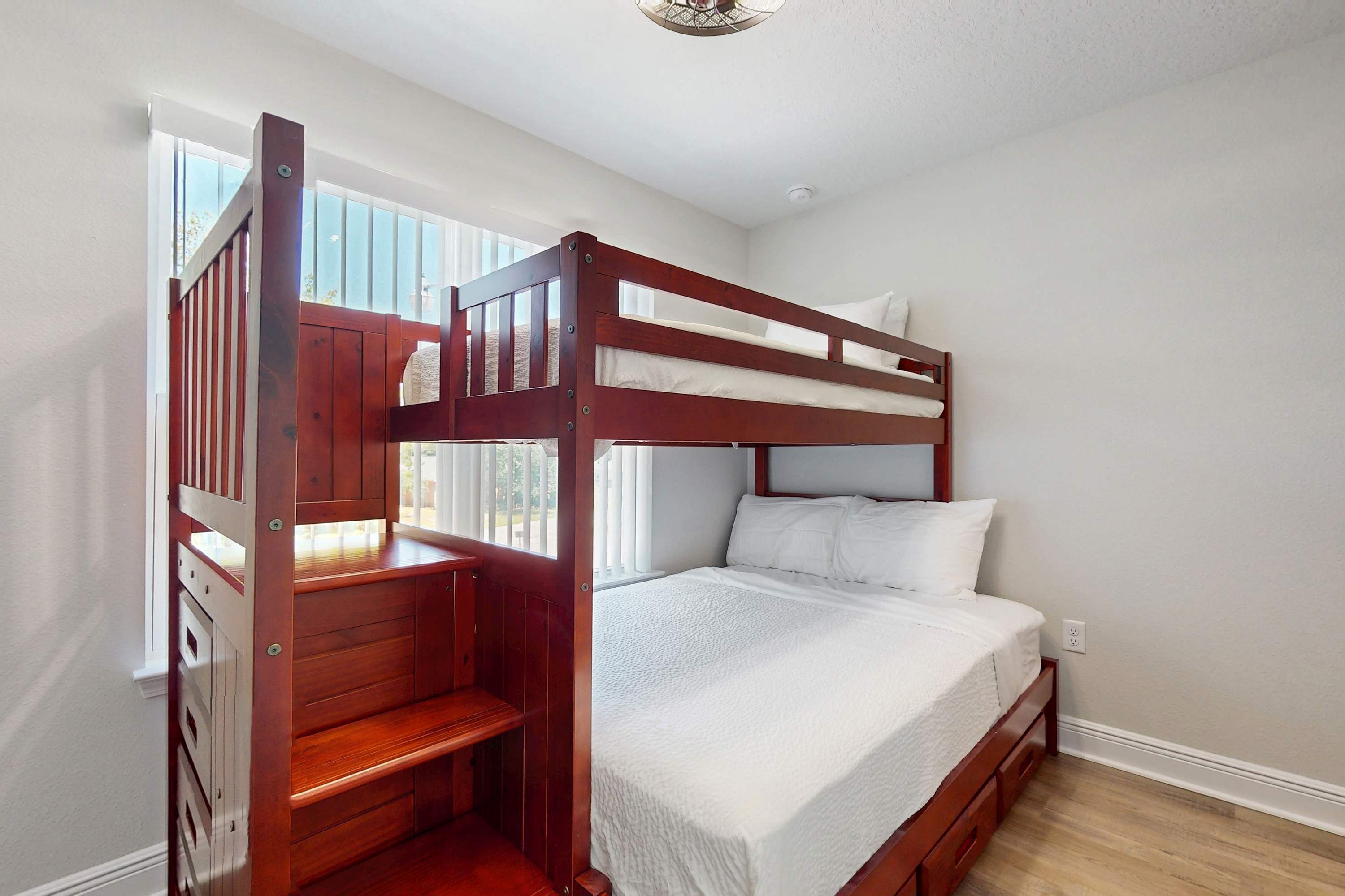 A bunk bed with a wooden frame and white bedding, a wooden desk, and a window with blinds.