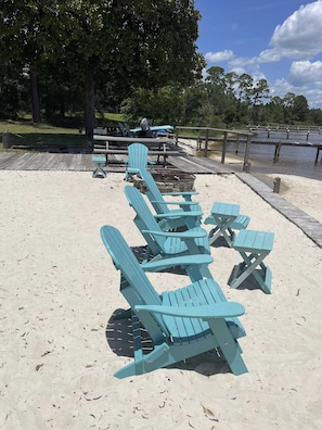 Ein Strandbereich mit Sand, mehreren blauen Adirondack-Stühlen und Picknicktischen, einer Holzterrasse und einem Holzzaun.
