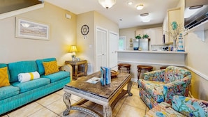 A living room with a teal sofa, a coffee table, and a colorful armchair. There is a kitchen in the background with white cabinets and a stove. - Bright townhome with patio, fenced yard & pool - near shops and dining (Miramar Beach)
