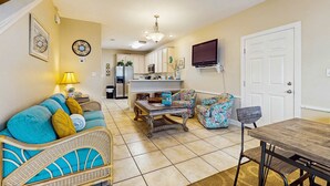A living room with a sofa, a coffee table, a dining table, and a TV mounted on the wall. - Bright townhome with patio, fenced yard & pool - near shops and dining (Miramar Beach)
