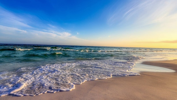 A beach with waves gently lapping onto the shore under a clear sky. - Bright townhome with patio, fenced yard & pool - near shops and dining (Miramar Beach)