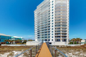 A tall, modern hotel with multiple balconies, a wooden boardwalk leading to the entrance, and a clear blue sky. - 9th Floor Gulf-Front Condo with Plenty Of Resort Amenities, Close To Dining (Navarre Beach)