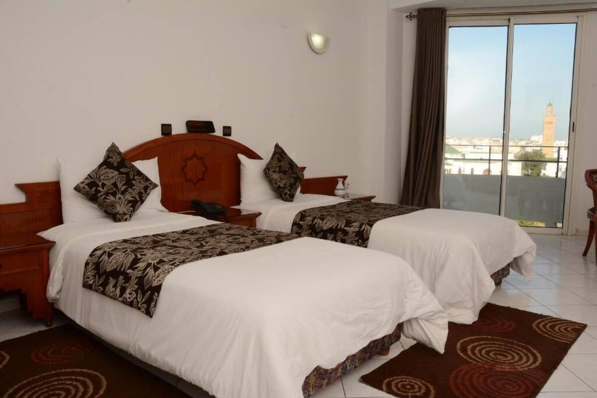 A hotel room with two beds, a wooden headboard, a nightstand, a small table, and a view of a cityscape through a large window.