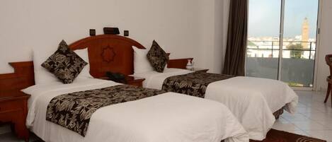 A hotel room with two beds, a wooden headboard, a nightstand, a small table, and a view of a cityscape through a large window.