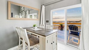Un coin repas avec une table en bois et des chaises blanches, un grand miroir et une vue sur la plage, visible à travers une porte-fenêtre coulissante.