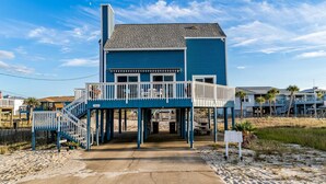 Une maison de deux étages située en bord de mer, dotée d’un balcon, d’une entrée couverte et d’une plage de sable en arrière-plan.