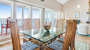 A spacious dining area with a glass table surrounded by wooden chairs, a bowl of fruit on the table, and a view of the ocean through large windows. - Gorgeous condo with ocean views, inviting balcony, bright interior, washer/dryer (Santa Rosa Beach)