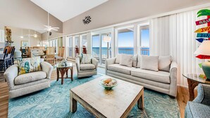 A spacious living room with a large window offering a view of the ocean, a cozy sofa, a coffee table with a bowl, and a dining area with chairs and a table. - Gorgeous condo with ocean views, inviting balcony, bright interior, washer/dryer (Santa Rosa Beach)