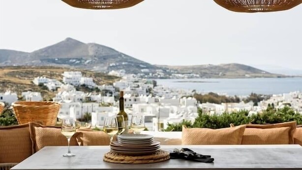 A table with a view of a coastal town, wicker chairs, and a bottle of wine. - Naousa Story 1 ❤ (Paros)