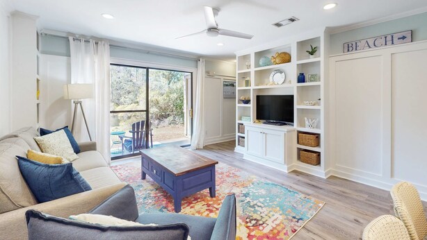 A spacious living room with a sofa, coffee table, and a television on a white cabinet. There is a ceiling fan, a large window with curtains, and a sign that says "BEACH". - Bright Townhouse with High-Speed WiFi, Private Washer&Dryer, and Pool (Santa Rosa Beach)