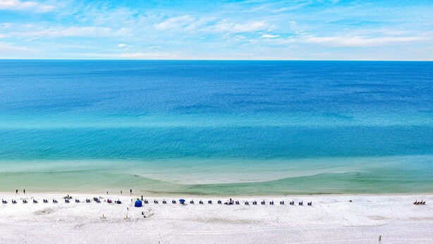 Ein Strand mit blauem Himmel, ruhiger See und sandigem Ufer, mit Liegestühlen und Sonnenschirmen.