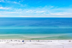 Uma praia com céu azul límpido, mar calmo e areal com cadeiras e guarda-sóis de praia.
